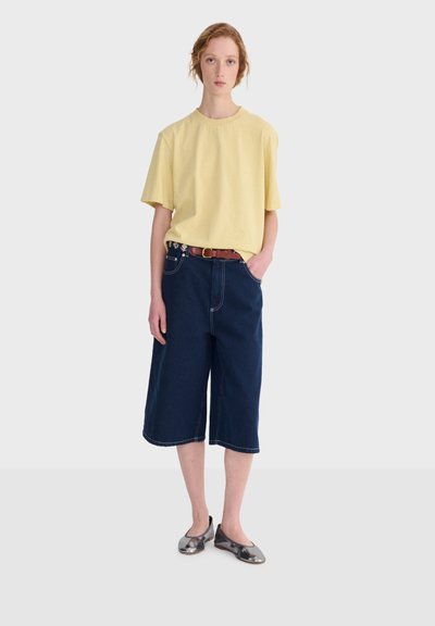 Jeune personne aux cheveux bouclés portant un t-shirt jaune ample, un short en jean bleu foncé arrivant aux genoux, une ceinture marron et des ballerines métalliques.