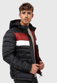 Jachetă puffer neagră cu dungi orizontale roșii și albe, fermoar frontal, glugă și textură netedă. Include manșete elastice și tiv cu șnur.