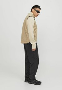 Beige ærmeløs veste over en cremefarvet langærmet top, sorte bukser og sorte sneakers. Model med solbriller, står sidelæns.