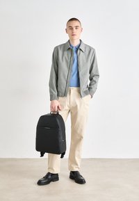 Jeune homme portant une veste grise, un polo bleu, un pantalon beige et des chaussures noires, tenant un sac à dos noir par sa poignée supérieure.