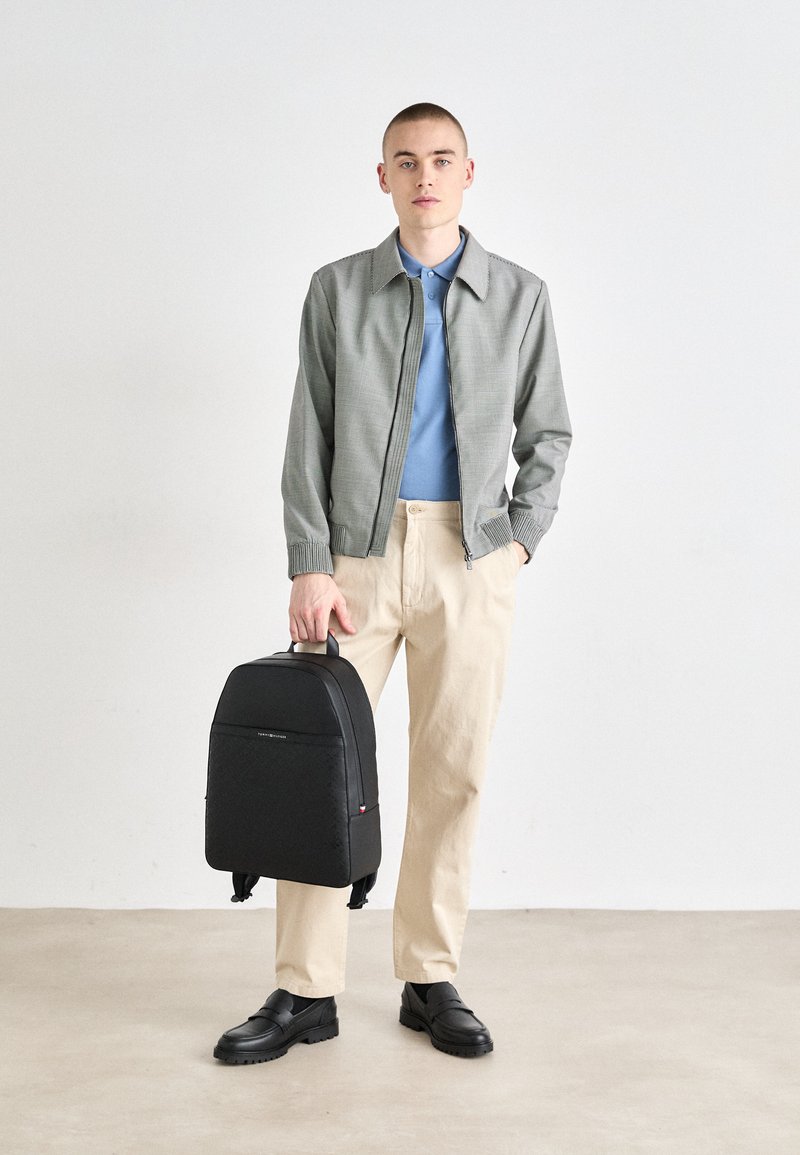 Jeune homme portant une veste grise, un polo bleu, un pantalon beige et des chaussures noires, tenant un sac à dos noir par sa poignée supérieure.