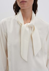 Blusa color crema con dettaglio di sciarpa annodata al collo, realizzata in tessuto liscio, con una vestibilità comoda e maniche lunghe.