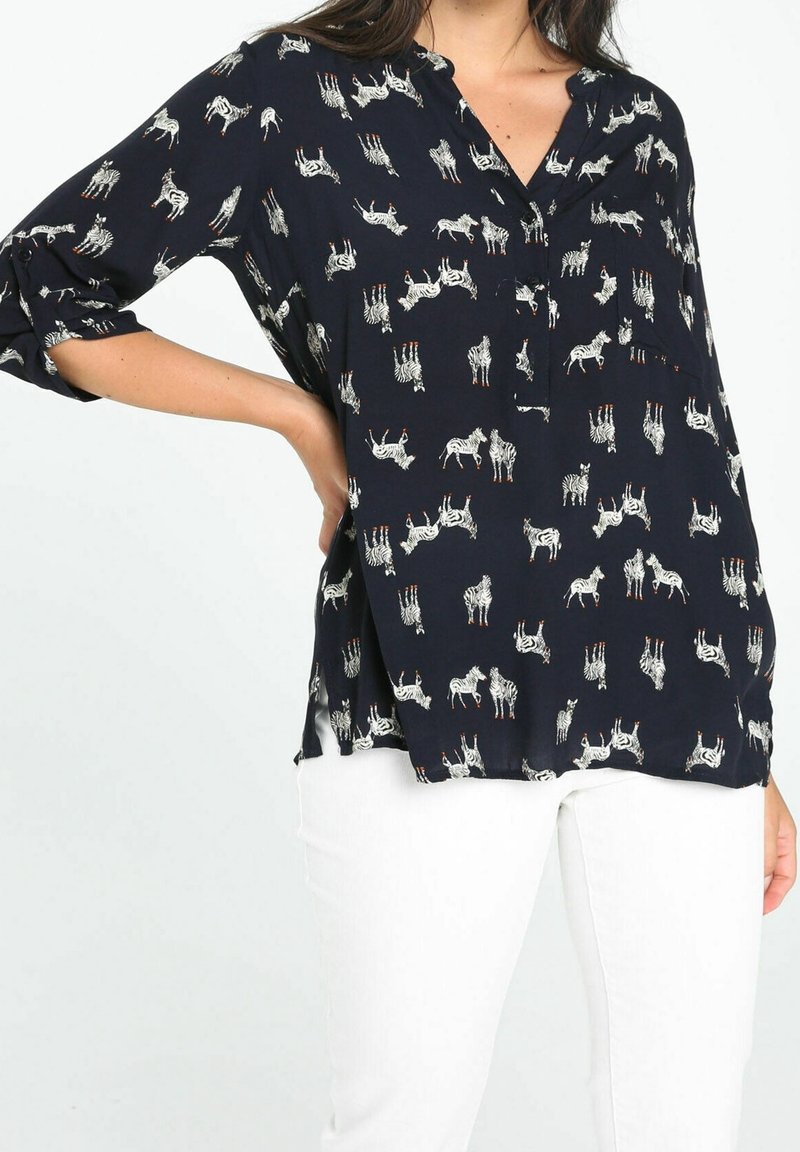 Blouse navy en tissu léger avec un imprimé zèbre intégral. Col boutonné, manches ¾ et fentes sur le côté à l'ourlet. Associée à un pantalon blanc.