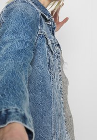 Džínová bunda ve světle modré s texturovaným povrchem. Má klasický límec, přední zapínání na knoflíky a dvě náprsní kapsy.