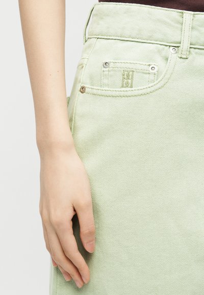 Gros plan de la main gauche d'une personne reposant à côté de la poche d'un pantalon en denim vert clair avec des rivets métalliques et des détails cousus.