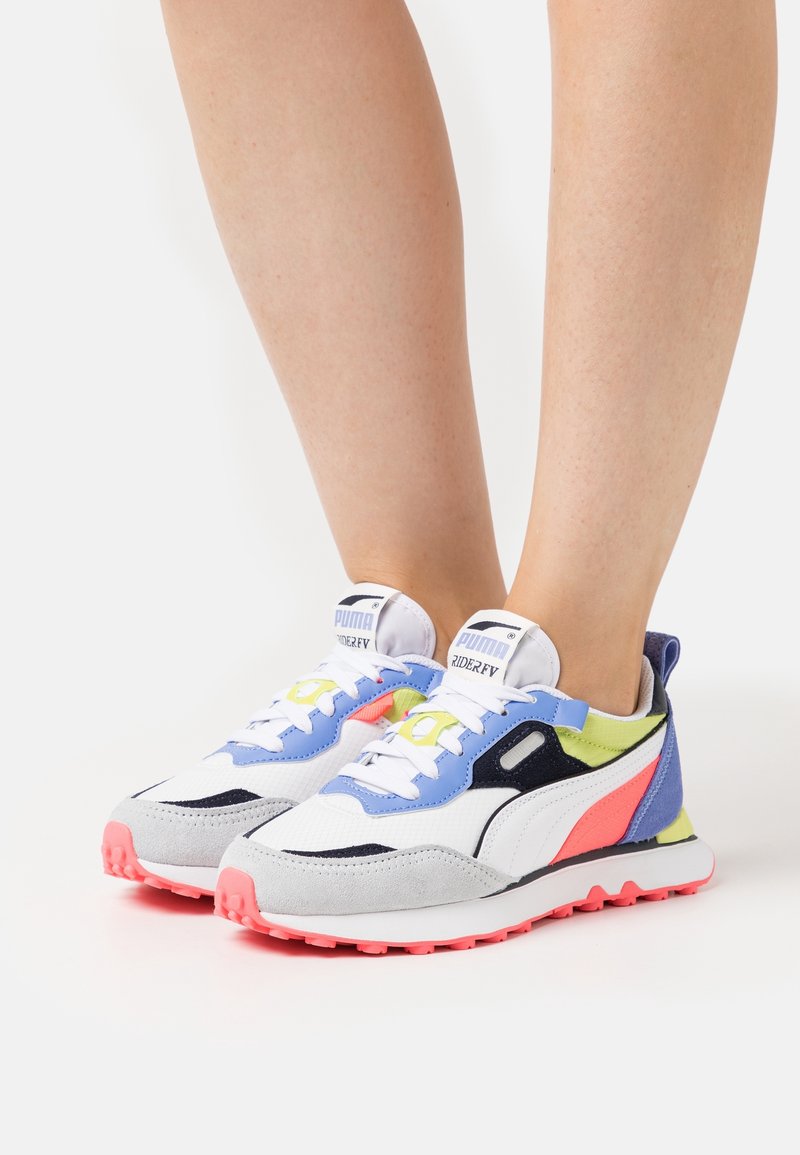 Personne portant des baskets Puma Rider FV blanches, bleues, jaune néon, noires et corail avec des semelles corail, debout sur une surface blanche.
