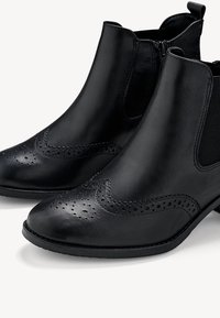 Bottes courtes en cuir noir avec détails brogue, panneaux élastiques latéraux et languettes de tirage à l'arrière, vues en angle trois-quarts avant.