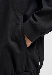 Schwarze Jacke aus glattem Material, mit elastischen Bündchen, Seitentaschen und einem gerafften Saum. Minimalistisches Design ohne sichtbare Logos oder Muster.