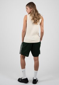 Femme aux longs cheveux ondulés portant un haut beige côtelé sans manches, un short rayé vert foncé avec des fentes latérales, des chaussettes blanches et des mocassins noirs, de dos.