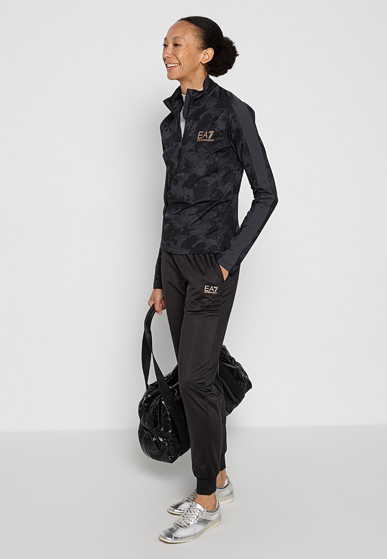 Černá sportovní bunda s vysokým límcem, maskáčový vzor, spárovaná s černými kalhotami s logem. Stříbrné tenisky, nese černou tašku.