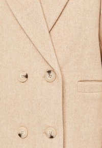 Blazer de mezcla de lana beige con doble botonadura, que presenta solapa de muesca, dos grandes botones redondos y un bolsillo en el pecho, con un acabado texturizado.