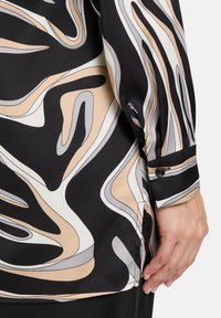Langärmlige Bluse aus schwarzem Stoff mit abstrakten Wirbeln in Beige, Grau und Creme; Knopfdetail am Bündchen, glatte Textur.