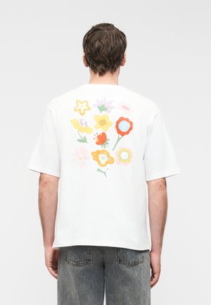 Άνδρας που φοράει λευκό κοντομάνικο πουκάμισο με πολύχρωμα αφηρημένα σχέδια λουλουδιών στην πλάτη, συνδυασμένο με ξεθωριασμένο γκρι τζιν παντελόνι.