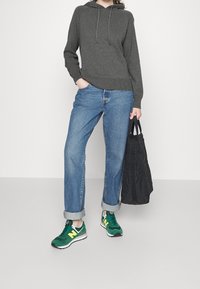 Sweat-shirt à capuche gris, jeans bleus retroussés, baskets vertes et jaunes, tenant un sac fourre-tout noir, sur un fond blanc uni.