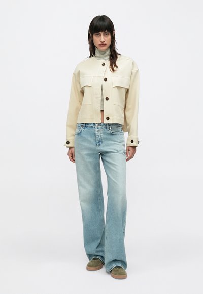 Γυναίκα με σκούρα μαλλιά φοράει μπεζ κοντό σακάκι πάνω από ριγέ ζιβάγκο, ανοιχτό μπλε τζιν παντελόνι φαρδιάς γραμμής και πράσινα σουέντ παπούτσια, στέκεται σε λευκό φόντο.