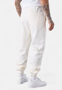 Mann mit tätowierten Armen, der weiße Jogginghosen und weiße Sneakers trägt, von hinten vor einem schlichten weißen Hintergrund gezeigt.