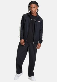 Zwarte zip-up jas met witte strepen langs de mouwen, gecombineerd met een zwart T-shirt en losse zwarte broek. Draagt witte sneakers.