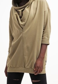 Sudadera oversize verde oliva con escote drapeado, mangas tres cuartos y detalles acanalados. Combinada con vaqueros negros rasgados.