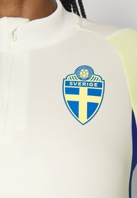 Top atletico bianco con colletto alto, caratterizzato da un logo a scudo blu e giallo etichettato "SVERIGE" sul petto; rifinito con bordi blu e gialli.