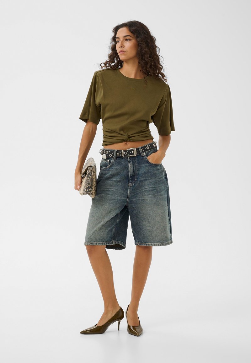 Femme aux cheveux bouclés portant un top vert olive à manches courtes, un short bermuda en jean taille haute, une ceinture cloutée, des talons olive et tenant une pochette en cuir de serpent.