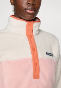 Primo piano di una persona che indossa una giacca in pile color-block con colletto alto e chiusura a bottoni corallo, caratterizzata dal logo Columbia sul petto.