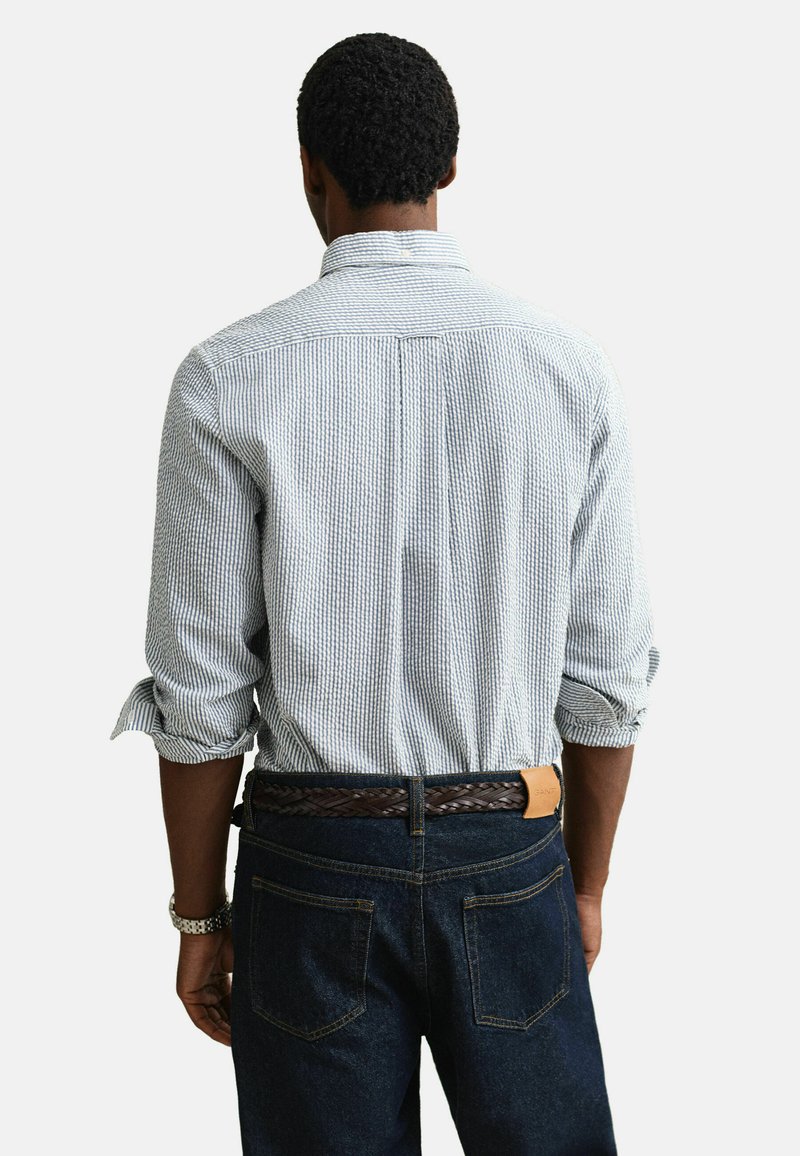 Hombre vistiendo una camisa de rayas azul claro con las mangas arremangadas, jeans azul oscuro y un cinturón marrón trenzado, visto desde atrás.