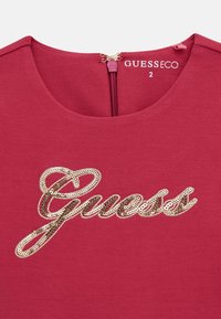 Haut en tissu rouge avec un décolleté en scoop, un logo "Guess" en sequins dorés et une fermeture éclair au dos avec une tirette dorée.