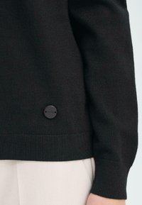 Maglione nero a maglia con orlo e polsini a coste, dotato di un dettaglio con logo nero tondo posizionato vicino alla parte inferiore sinistra dell'indumento.