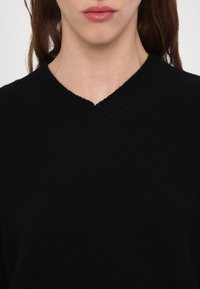 Partie inférieure du visage et cou d'une personne portant un pull en maille noire avec un col en V, lèvres fermées et cheveux bruns visibles.