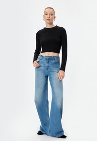 Zwarte, korte gebreide trui met ribbelstructuur en een ronde halslijn, gecombineerd met lichtblauwe wijde jeans. Schoeisel niet zichtbaar.
