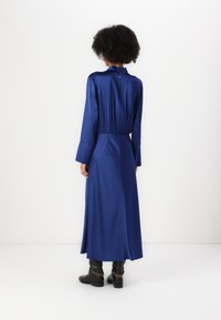 Abito lungo in satin blu con colletto alto, maniche lunghe, vita arricciata e dettaglio plissettato sul retro, abbinato a stivaletti neri alla caviglia.