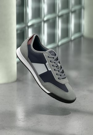 Baskets gris et bleu marine avec semelle blanche et logo "BOSS", flottant au-dessus d'un sol en béton poli près d'un mur en verre dépoli.