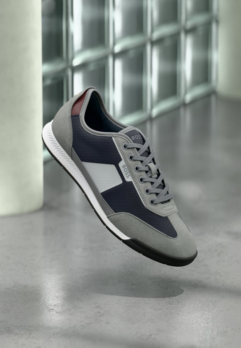 Γκρι και ναυτικό μπλε αθλητικό παπούτσι με λευκή σόλα και το λογότυπο "BOSS", αιωρείται πάνω από το γυαλισμένο τσιμεντένιο δάπεδο κοντά σε θολωμένο γυάλινο τοίχο.
