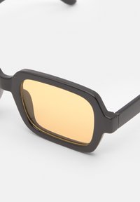 Lunettes de soleil rectangulaires noires avec des verres teintés jaunes, présentant une monture épaisse et une texture subtile. Comprend des branches droites.