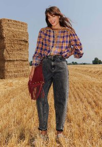 Chemise à carreaux violette, jaune et blanche ; jeans en denim noir taille haute ; sac à bandoulière rouge à franges. Décor rural avec des bottes de foin.