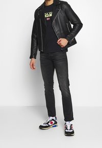 Chaqueta de cuero negra de motorista con cremalleras plateadas, usada sobre una camiseta azul marino con texto en neón. Combinada con unos jeans oscuros y zapatillas multicolores.