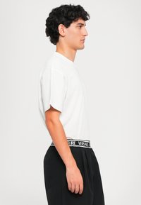 Jovem com cabelo encaracolado e escuro está de perfil, vestindo uma t-shirt branca e calças pretas com um cós visível com o logótipo da Versace.