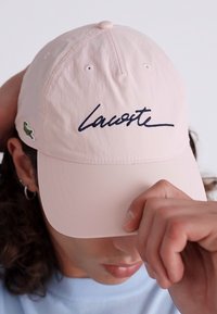 Svijetloružičasti baseball kapica s tamnoplavim kurzivnim logotipom "Lacoste" i zelenim logotipom krokodila sa strane, s odijeljenom krova i podesivim remenom.