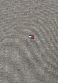 Grau strukturiertes Gewebe mit einem kleinen roten, weißen und blauen gestickten Logo auf der linken Seite. Das Material hat eine gerippte Optik.