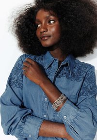 Chemise en denim bleu clair avec des détails brodés complexes sur les épaules, des manches longues et un devant à bouton. Bracelet en argent au poignet.
