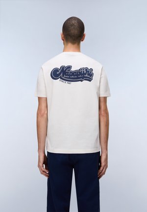 Fehér rövid ujjú inget viselő férfi, amelyen hátul tengerészkék "Napapijri Since 1987" logó található, sötétkék nadrággal, háttal állva.