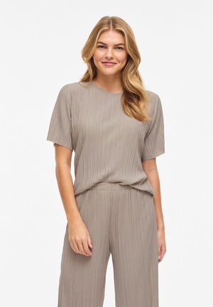 Femeie cu păr lung, blond, purtând un top taupe plisat cu mâneci scurte și pantaloni largi, zâmbind pe un fundal alb simplu.