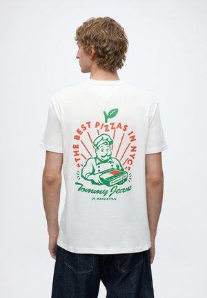 Personne aux cheveux courts et bouclés portant un t-shirt blanc avec un chef pizza rétro et le texte "The Best Pizzas in NYC" dans le dos.