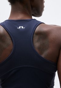 Αθλητικό άτομο εμφανιζόμενο από πίσω που φοράει μπλε σκούρο τοπ χωρίς μανίκια με λευκό μικρό λογότυπο κοντά στον λαιμό, πάνω σε απλό φόντο.