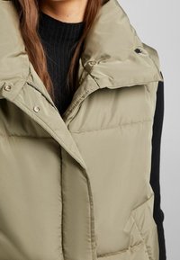 Prošívaná vesta v tlumené zelené barvě s vysokým límcem, patentními knoflíky a bočními kapsami, doplněná černým žebrovaným svetrem pod ní.