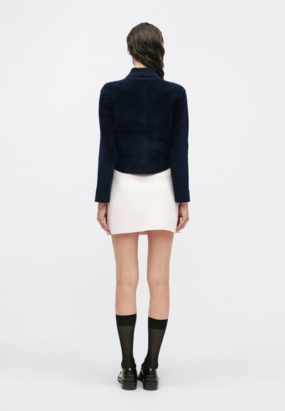 Mujer vistiendo una chaqueta corta azul marino, una minifalda blanca, medias altas de red negras y mocasines negros, de pie de espaldas sobre un fondo blanco.