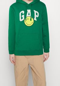 Sudadera con capucha verde con un bolsillo delantero, que presenta un gráfico de una cara sonriente y el texto "GAP" en blanco encima. Material de algodón con una textura suave.