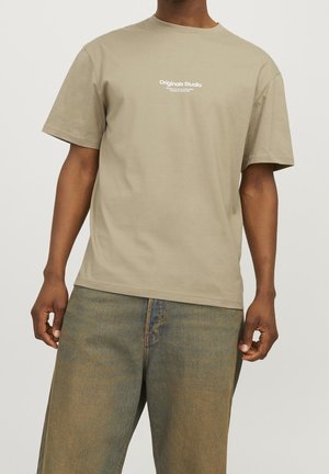 Homme portant un t-shirt beige à manches courtes avec le texte "Originals Studio" et un jean bleu délavé, debout devant un fond clair uni.