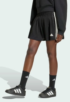 Person iført sort plisseret sportnederdel, sorte høje sokker med hvide logoer og sorte sneakers med hvide striber.