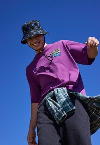 Jeune personne portant un chapeau seau fleuri, un t-shirt violet Venice Beach et une chemise à carreaux nouée autour de la taille, souriant devant un ciel bleu clair.